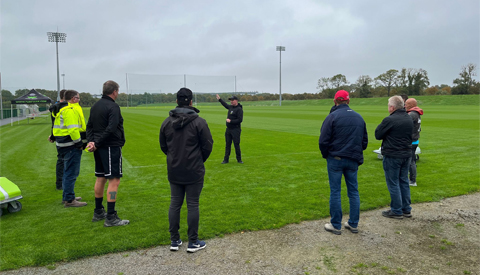 Nine guys standing in a half circle looking at a guy who's talking at a Turf Tank field day